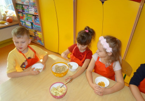 Troje dzieci siedzi przy stole, dziewczynka w środku nakłada łyżką do swojej miski płatki owsiane.